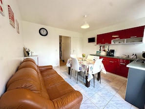 Un salon avec un canapé en cuir marron, une table à manger avec des chaises, une cuisine avec des placards rouges et une horloge au mur.