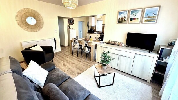 Ein modernes Wohnzimmer mit einer Couch, einem Couchtisch und einem Fernseher. Im Hintergrund befindet sich eine Küche mit Essbereich.