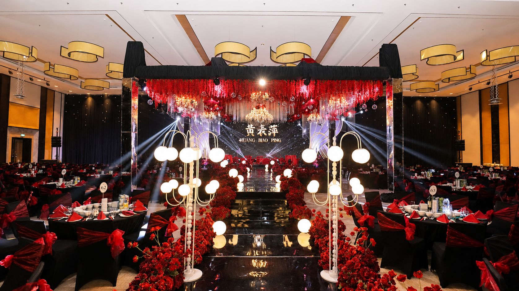 Un salón de banquetes con mesas dispuestas para un evento formal, que cuenta con un escenario con fondo rojo y dorado y un candelabro.
