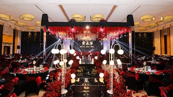 Un salón de banquetes con mesas dispuestas para un evento formal, que cuenta con un escenario con fondo rojo y dorado y un candelabro.