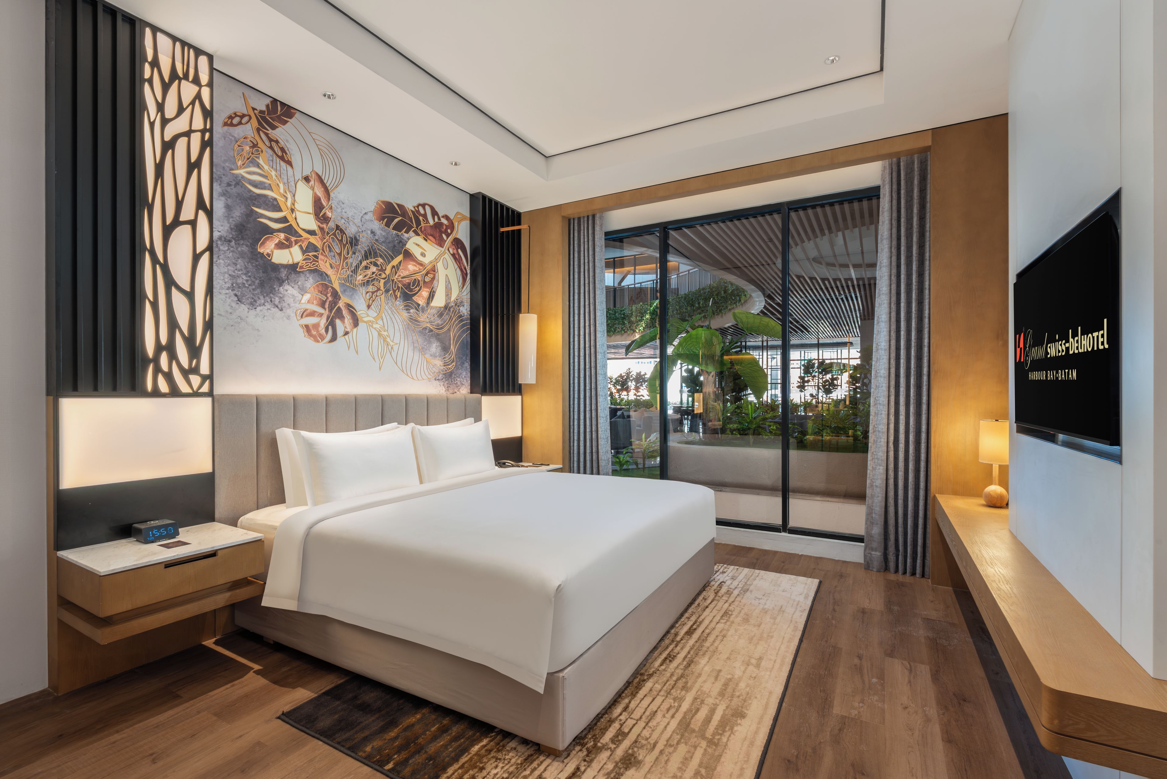 A modern hotel room with a large bed, a nightstand, a digital clock, a wall-mounted TV, and a large window with a view of greenery.