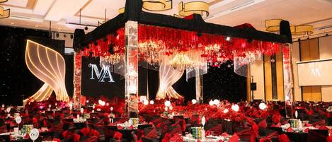 Un salón de banquetes con mesas dispuestas para un evento formal, con un escenario que incluye un telón de fondo y una gran pantalla que muestra las iniciales 'MA'.