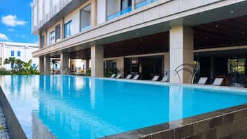 A swimming pool with lounge chairs and a covered area, surrounded by a building with multiple floors and large windows.