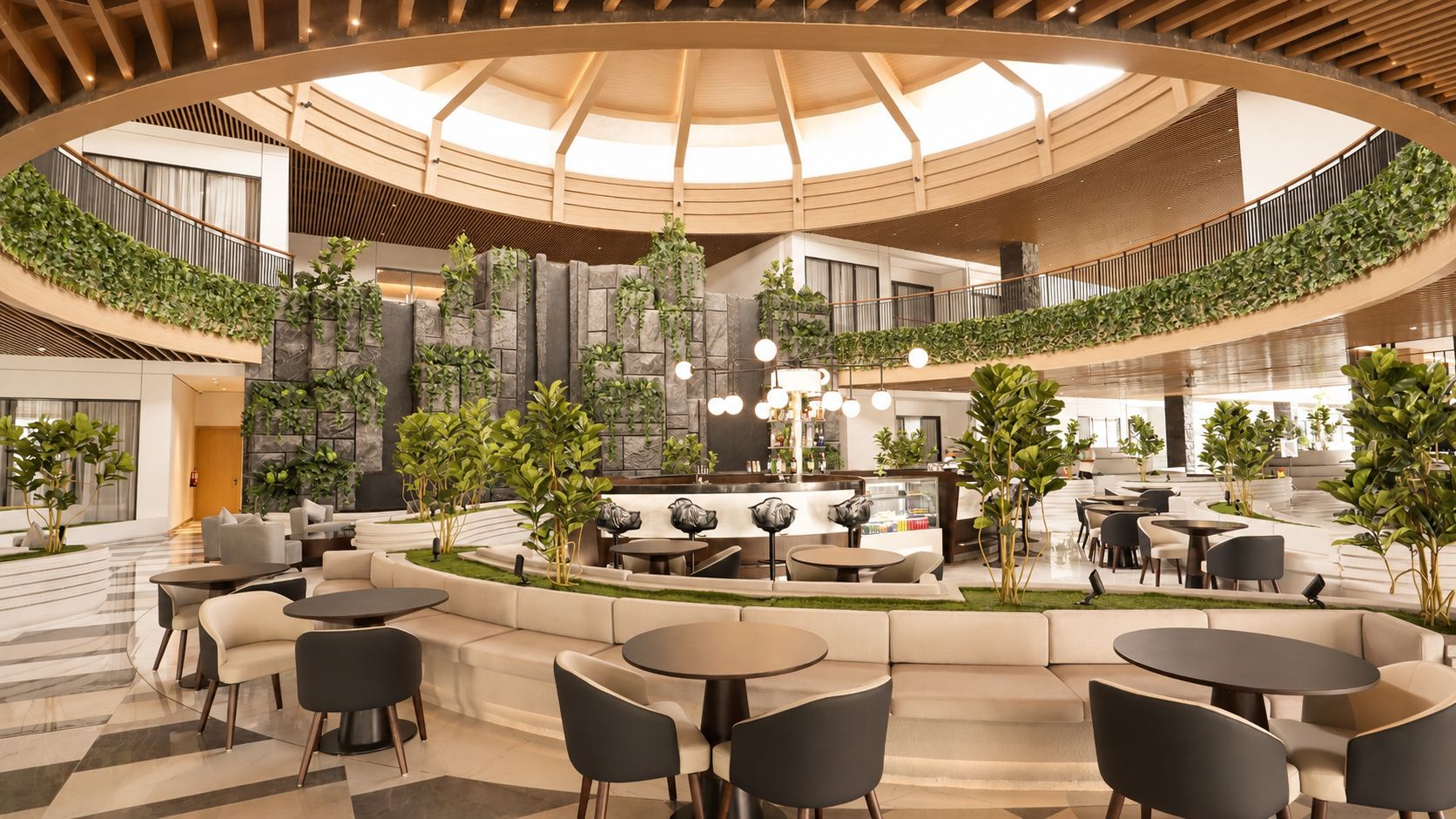 A spacious indoor area with multiple seating arrangements, including sofas and tables, surrounded by greenery and a central bar area.