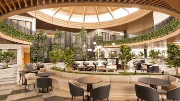 A spacious indoor area with multiple seating arrangements, including sofas and tables, surrounded by greenery and a central bar area.