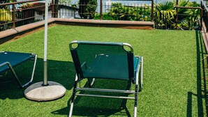 Een terras met kunstgras, een groene lounge stoel, een witte geribbelde parasol en uitzicht op een meer en bergen.