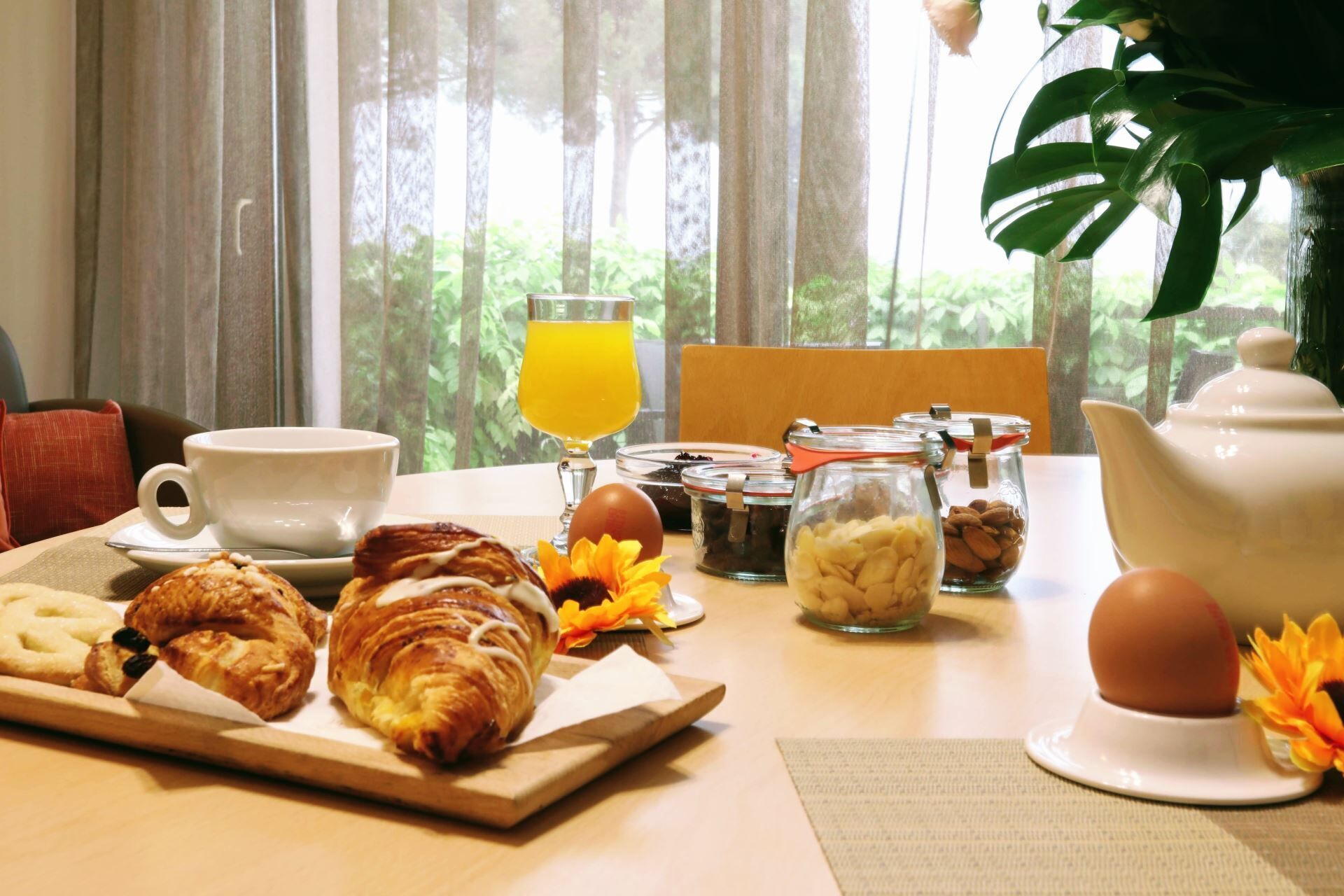 A breakfast setting with croissants, eggs, and a fruit drink on a table with a view of trees through sheer curtains.