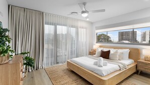 A modern bedroom with a large bed, a ceiling fan, sheer curtains, and a view of buildings through the window.