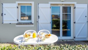 Eine Terrasse mit einem für das Frühstück gedeckten Tisch, zwei Fenster mit Rollläden und eine Tür mit Glasfeldern.