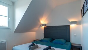Ein Schlafzimmer mit schrägverdeckter Decke, einem Bett mit türkisfarbenem Bettzeug, zwei Nachttischlampen und einem Fenster.