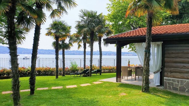 A wooden cabin with a stone exterior, a patio with outdoor furniture, and a view of a lake with palm trees in the foreground.