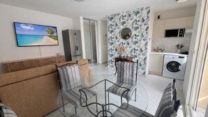 Ein modernes Wohnzimmer mit einem Glastisch, gestreiften Sesseln, einem beige-farbenen Sofa, einem Flachbildfernseher und einer Küche mit Waschmaschine.