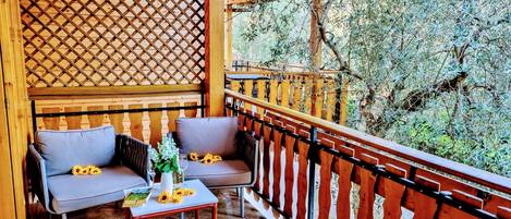 A balcony with a wooden lattice wall, two grey cushioned chairs, a small table with a vase of flowers, and a view of trees.