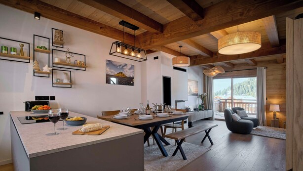 A modern kitchen with a central island, dining area with a wooden table, and a cozy seating area with a chair and lamp.