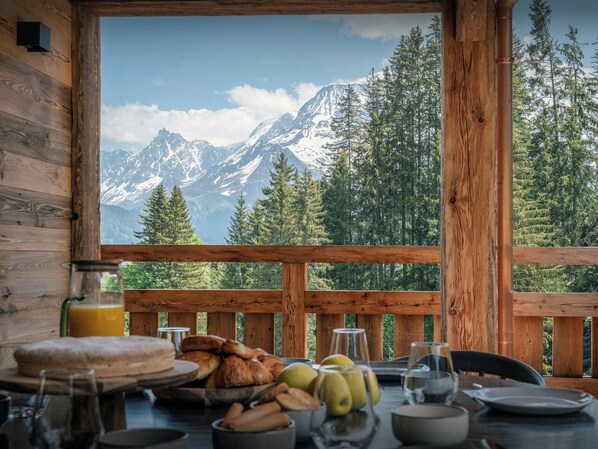Eine Holzhütte mit Frühstückstisch, gedeckt mit Gebäck, Früchten und Gläsern, mit Blick auf eine gebirgige Landschaft.