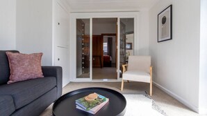 Un salon avec un canapé foncé, un fauteuil clair, une table basse avec des livres et une porte-fenêtre coulissante donnant sur une chambre.