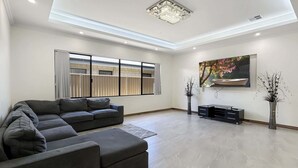 A modern living room with a sectional sofa, a flat-screen TV, and a large window with blinds. - Spacious 4 bed home with open plan living (Southern River)