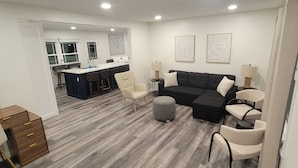 A modern living room with a black sectional sofa, two armchairs, and a small ottoman. A kitchen area is visible in the background with a dining table and chairs.