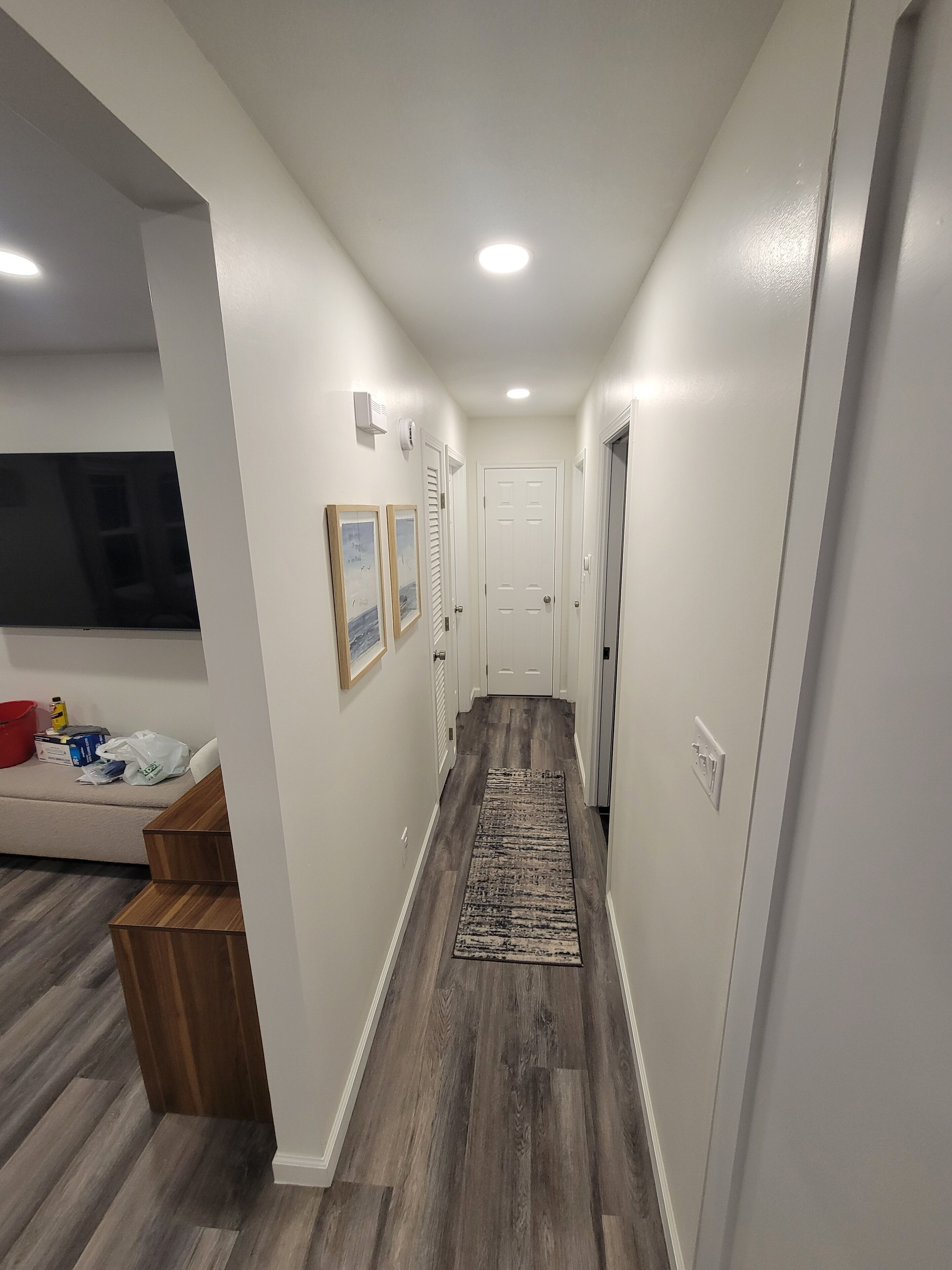 A narrow hallway with wooden flooring, white walls, and a central rug. There are two doors, one with a handle and one with a keyhole. A small trash bin is visible on the left side.