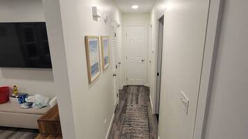 A narrow hallway with wooden flooring, white walls, and a central rug. There are two doors, one with a handle and one with a keyhole. A small trash bin is visible on the left side.