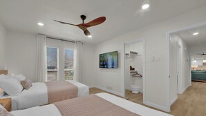A modern hotel room with two beds, a ceiling fan, a TV, and a view of the kitchen area.