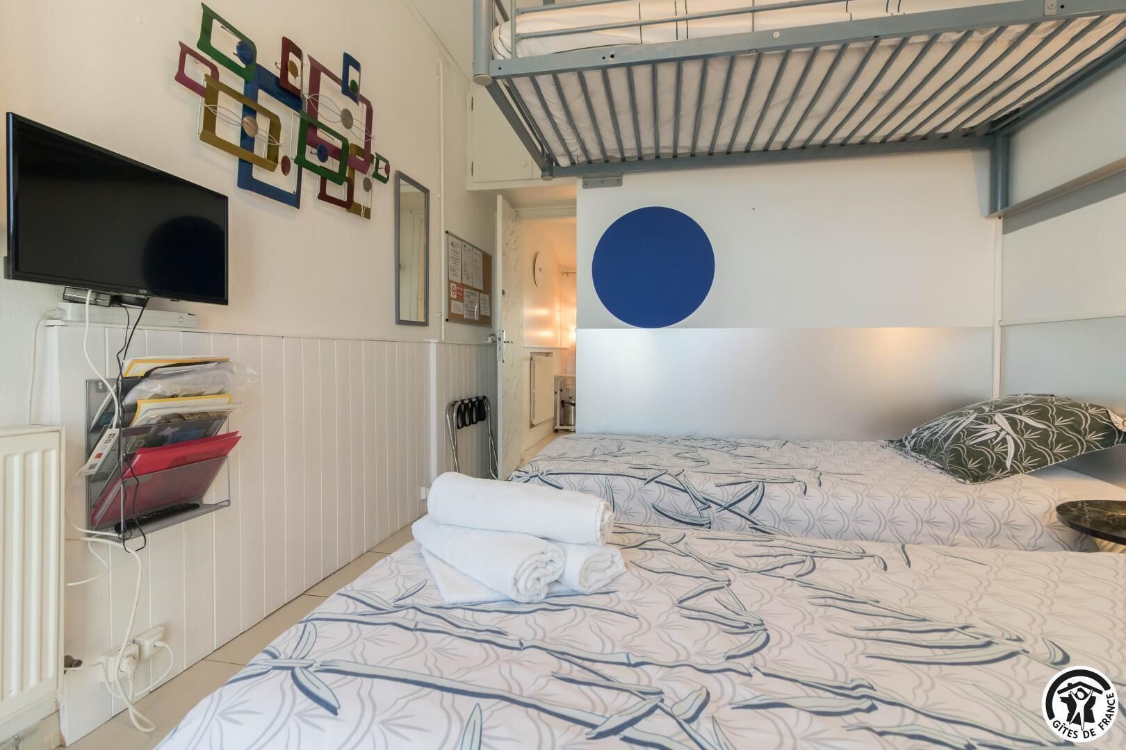 Une chambre d’hôtel avec deux lits, un téléviseur à écran plat, une étagère fixée au mur avec des classeurs, et un grand cercle bleu peint sur le mur.