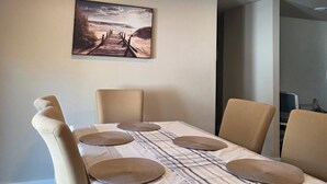 Un coin repas avec une table dressée pour quatre personnes, des chaises de couleur beige et un tableau représentant une scène de plage accroché au mur.
