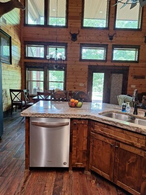 A kitchen with a central island, a dishwasher, wooden cabinets, and a large window overlooking greenery. - Serenity, secluded 4-bedroom, 3 bath spacious lux cabin with lots of amenities (Ringold)
