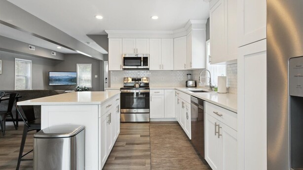 A modern kitchen with white cabinetry, stainless steel appliances, a central island, and a dining area with a TV. - Beach Bliss in Hampton – Minutes to the Shore! (Hampton)