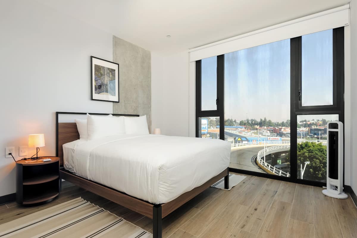 A modern hotel room with a large bed, a nightstand, a lamp, a framed picture on the wall, and a large window with a city view.