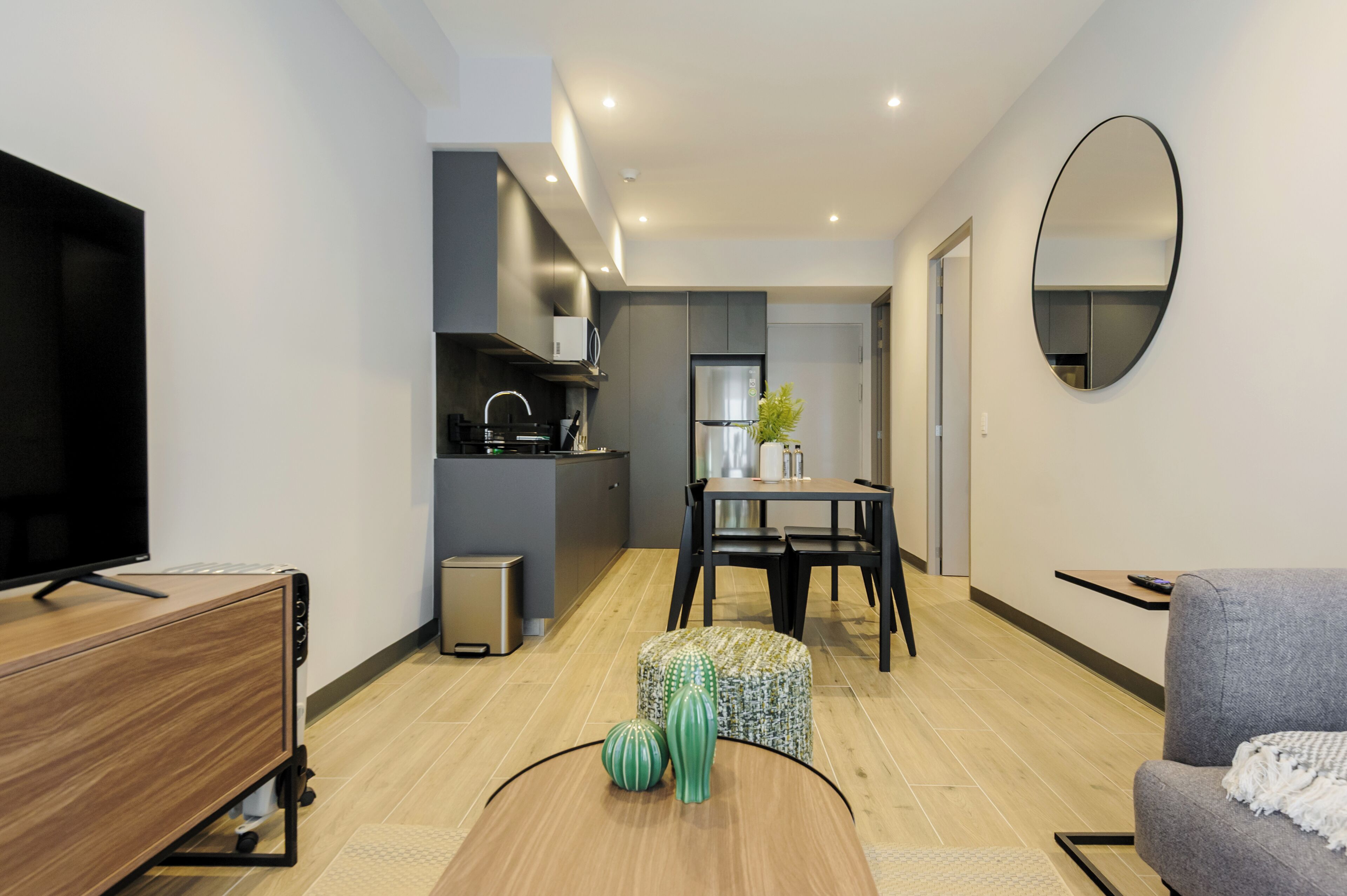 Un salon moderne doté d’un téléviseur à écran plat, d’un meuble en bois, d’un coin repas avec une table et des chaises, et d’une cuisine équipée d’un four à micro-ondes et d’un réfrigérateur.