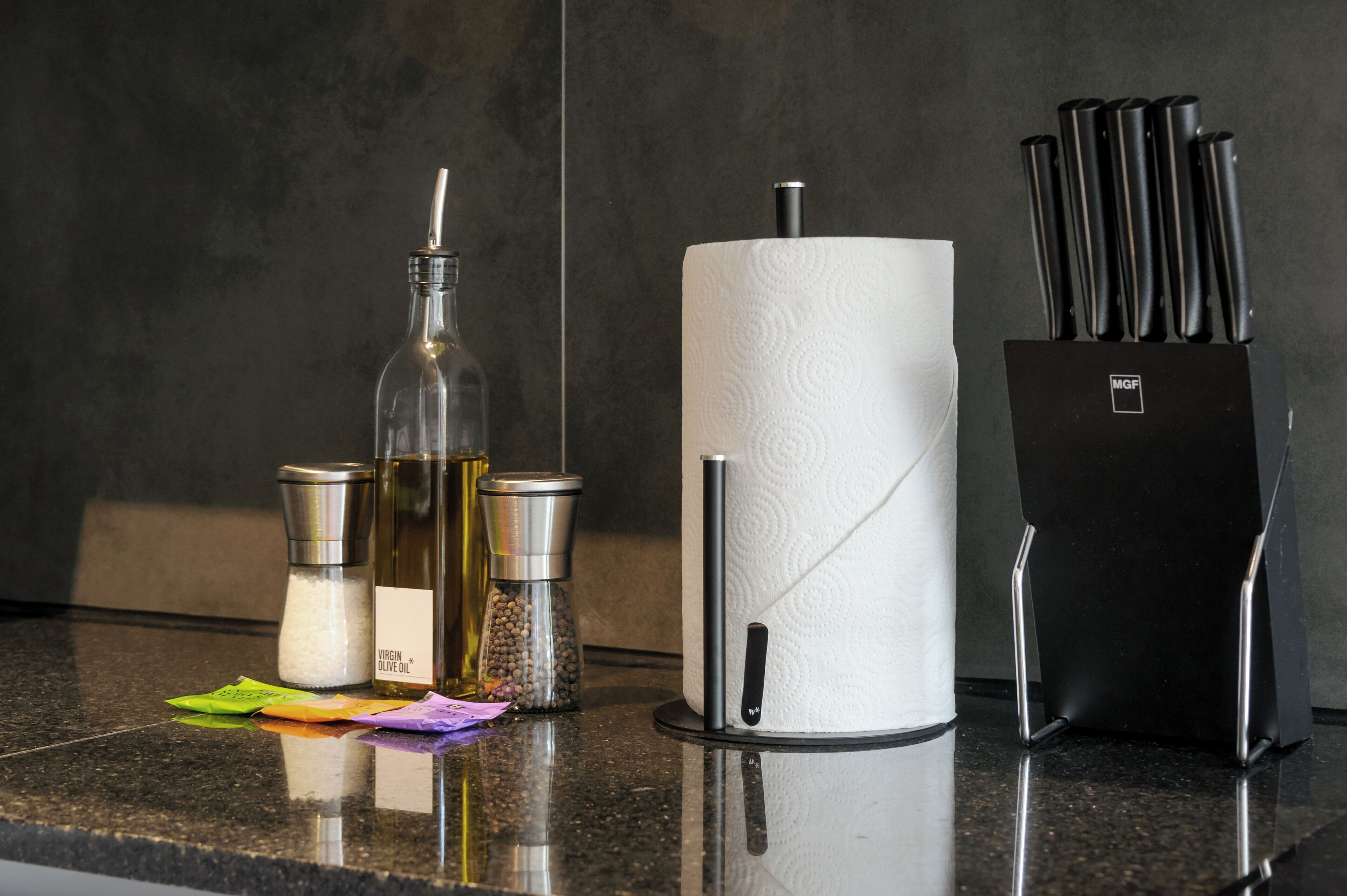 A kitchen counter with a paper towel holder, a salt and pepper shaker, a bottle of olive oil, and a knife block.