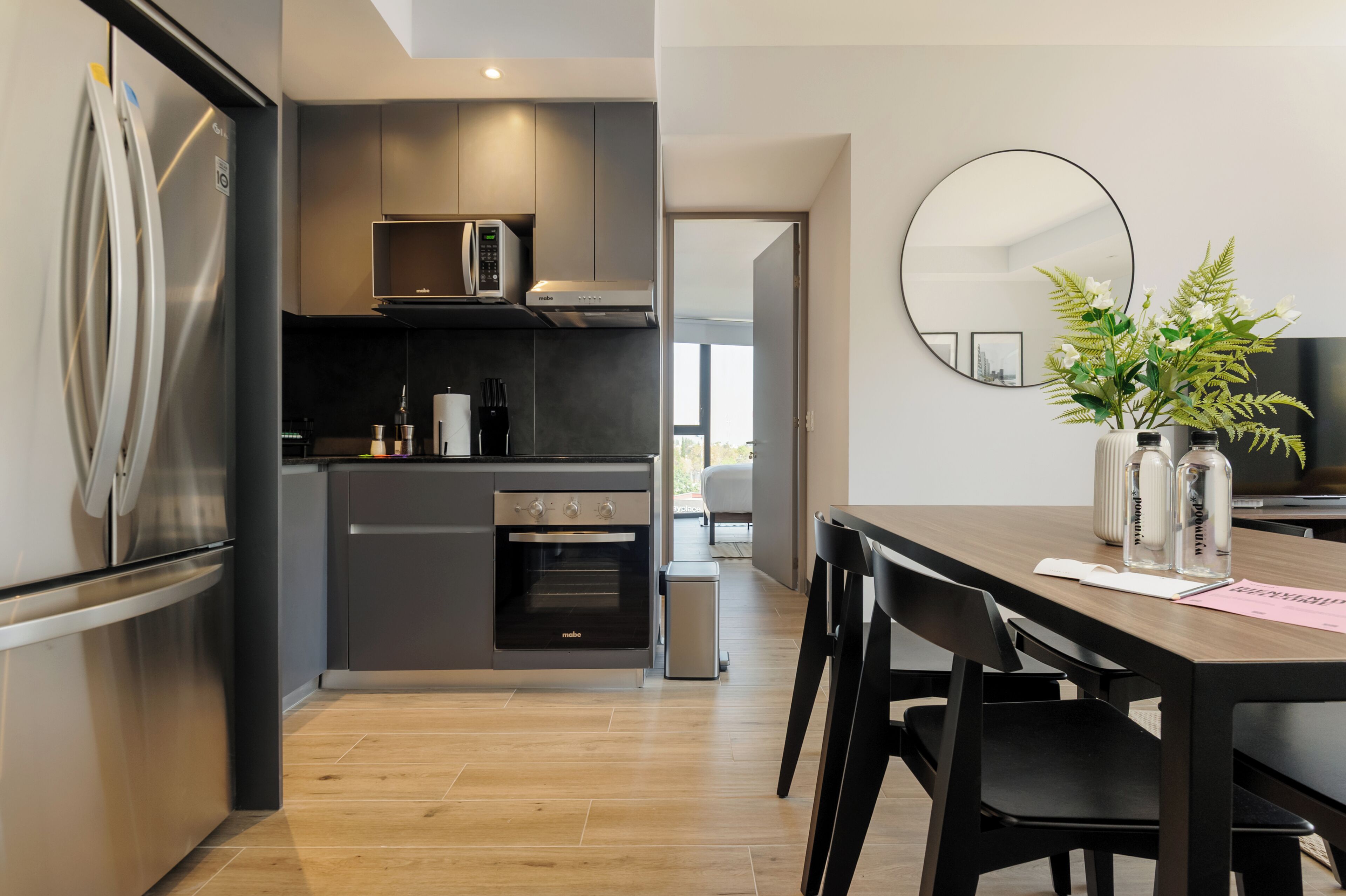 A modern kitchen with a built-in oven, microwave, and stainless steel refrigerator. There is a dining table with chairs and a vase with flowers.