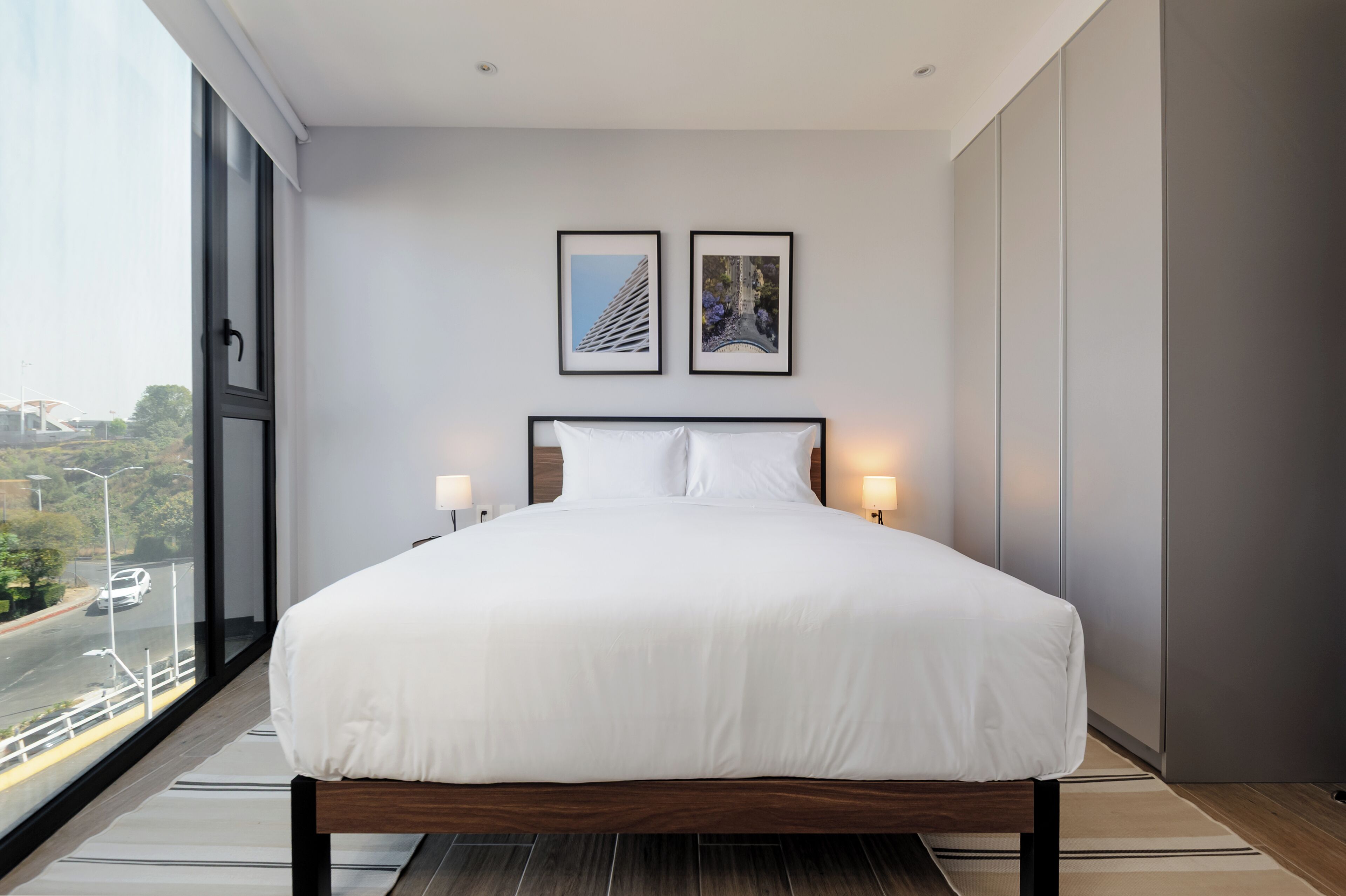 A modern bedroom with a large bed, two framed pictures on the wall, and a city view through the window.