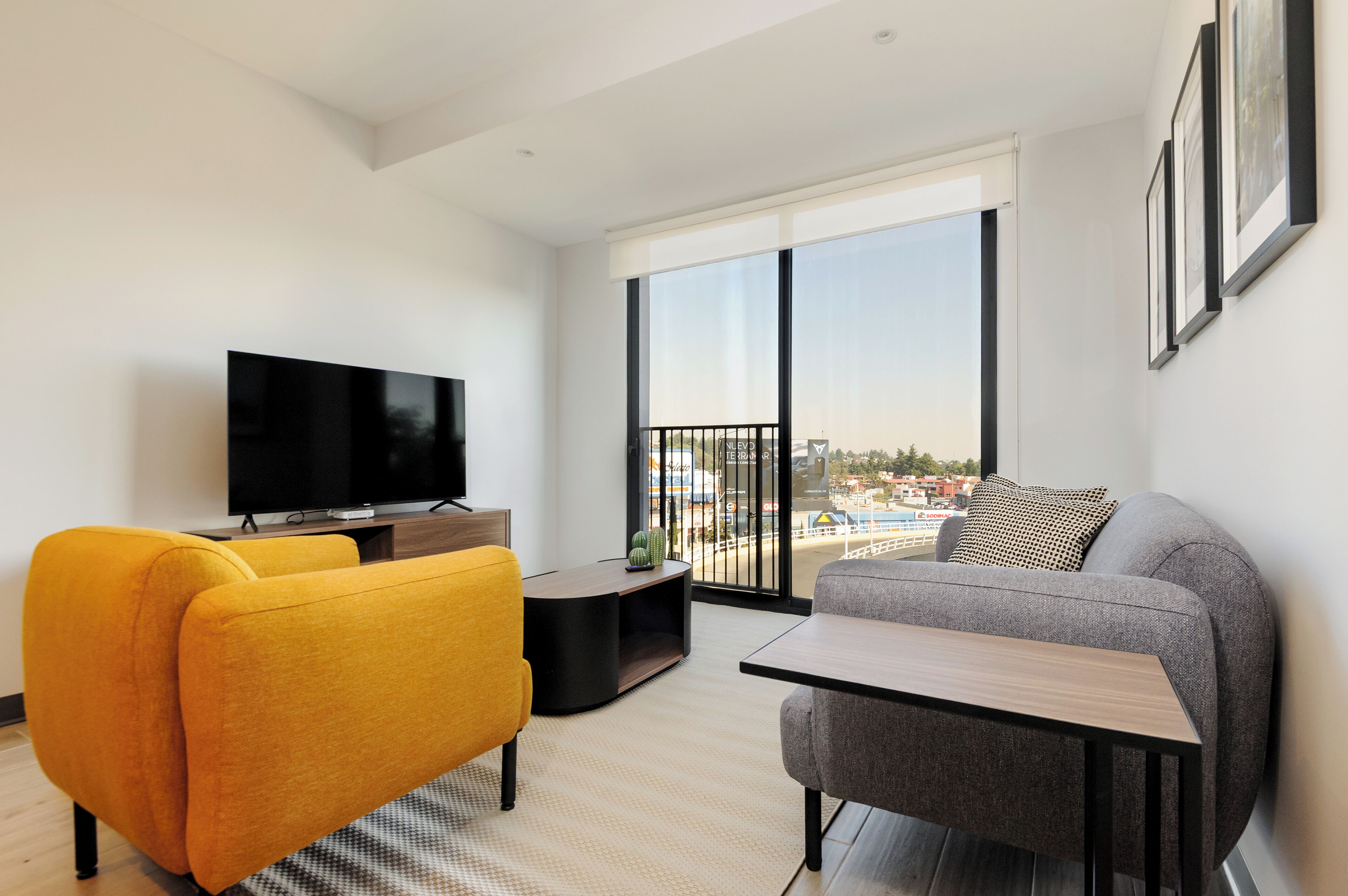 A modern living room with a large TV, a sofa, a yellow armchair, and a balcony with a city view.