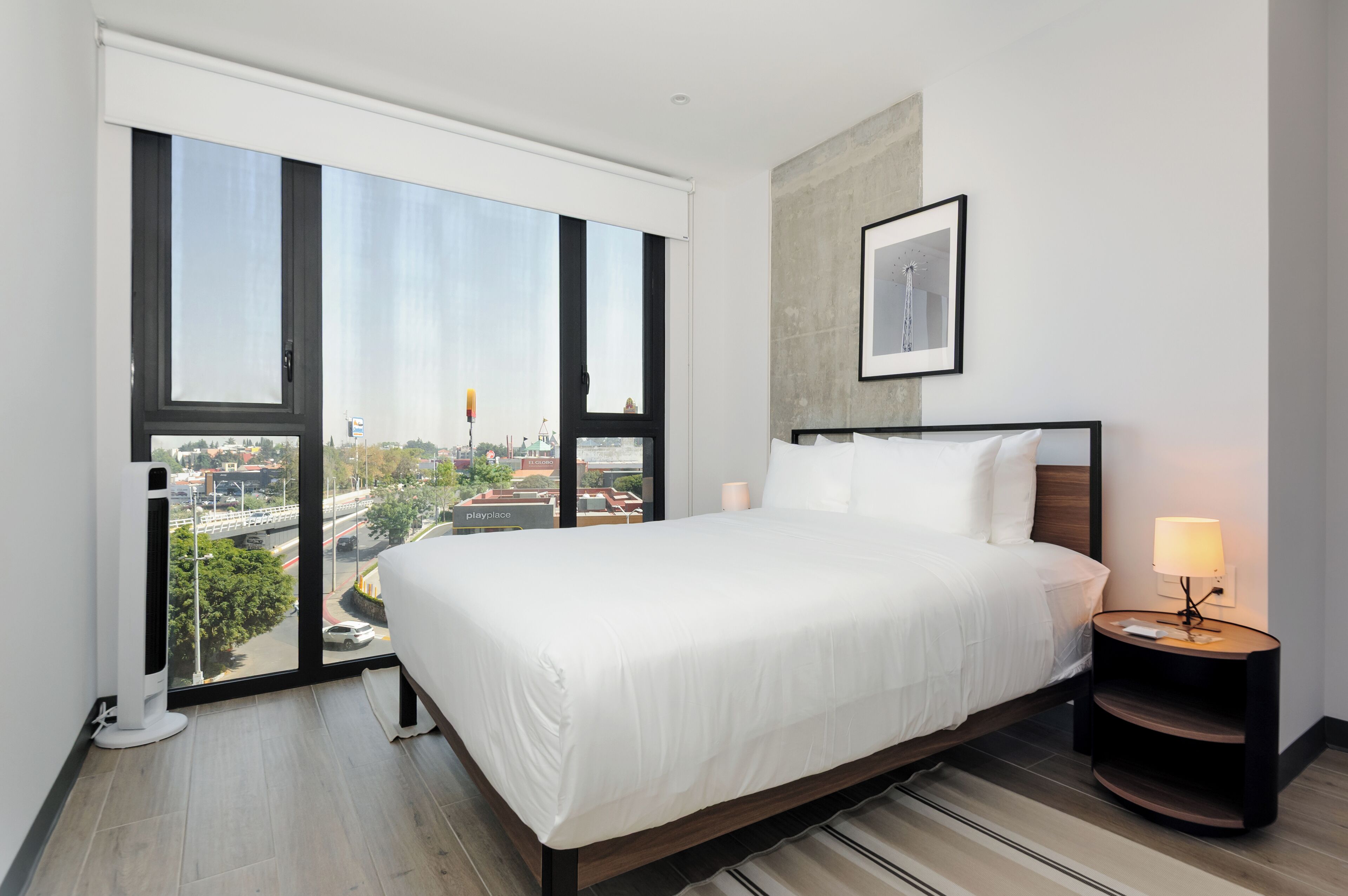 A hotel room with a large bed, a nightstand with a lamp, and a view of the city through floor-to-ceiling windows.