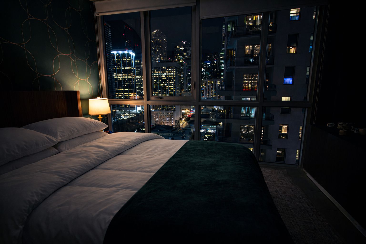 A bedroom with a large bed, a nightstand, a lamp, and a view of a cityscape at night.
