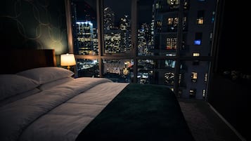A bedroom with a large bed, a nightstand, a lamp, and a view of a cityscape at night.