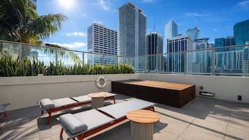 A rooftop terrace with lounge chairs, a wooden bench, and a view of the city skyline.