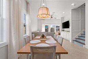 A modern dining room with a wooden table, white chairs, and a large chandelier. - Steps to Gulf | 4BR w/ Private Pool, Putting Green, Fire Pit & Golf Simulator – Sleeps 14 (Panama City Beach)