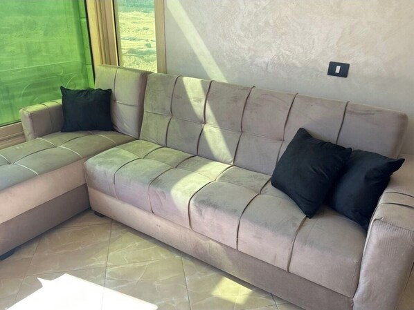 Ein modulares Sofa mit heller Stoffbezüge und dunklen Kissen, platziert in einem Raum mit großem Fenster und Lichtschalter an der Wand.
