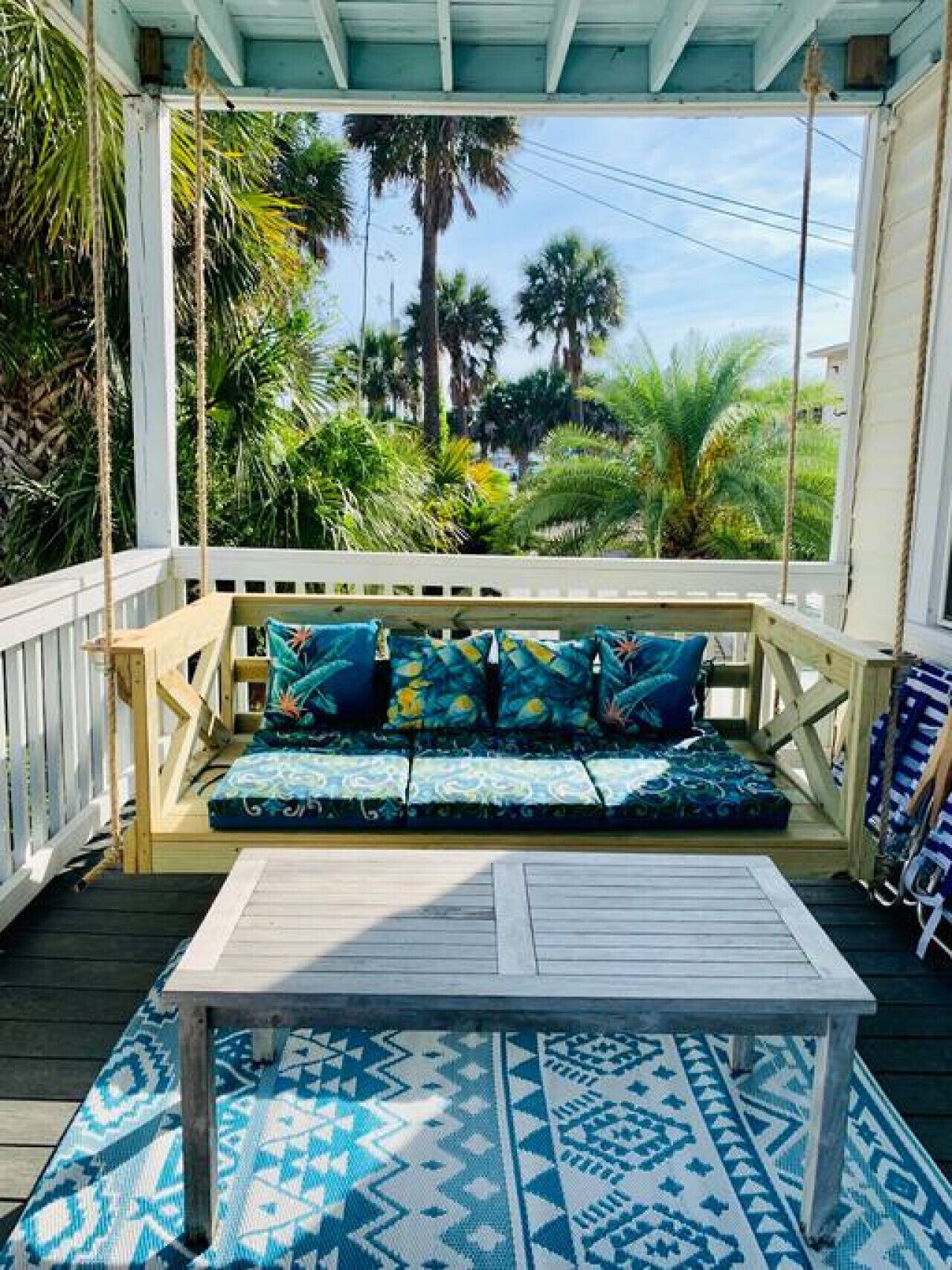 A wooden deck with a swing chair, a cushioned bench with patterned cushions, and a patterned rug.
