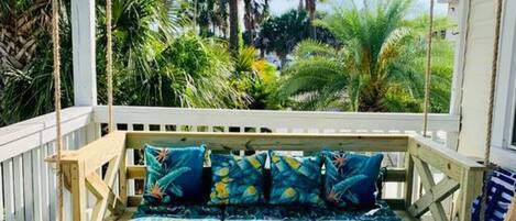 A wooden deck with a swing chair, a cushioned bench with patterned cushions, and a patterned rug.