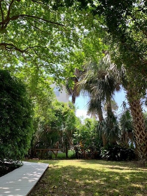 A garden with a paved walkway, various trees, and a building in the background. - “Sea Spot - Down!” Ideal Location & Beach Access! (Folly Beach)