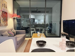 Ein modernes Wohnzimmer mit einer Couch, einem Esstisch mit weißem Tischlein und einem Fernseher auf einem Schrank.