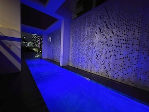 Ein Poolbereich mit blau beleuchtetem Boden, einer dekorativen Lochwand und Blick auf eine nächtliche Stadtlandschaft.