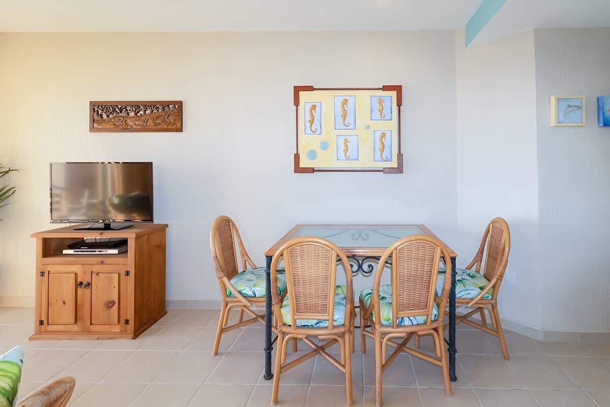 Un espace repas comprenant une table et des chaises, un téléviseur posé sur un meuble en bois, et des œuvres d’art murales.
