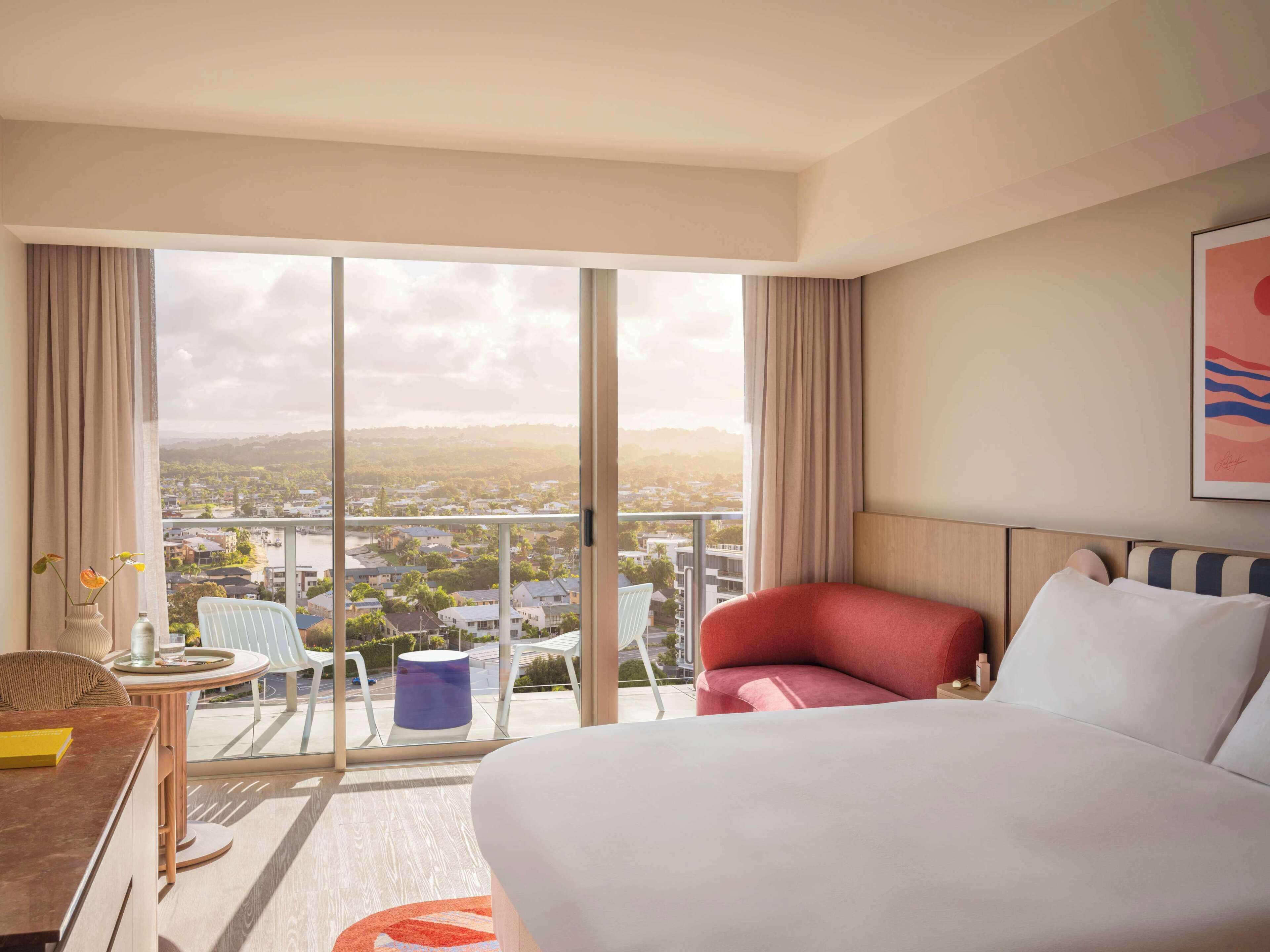 A hotel room with a large bed, a desk with a chair, a small table with a vase, and a view of a cityscape through large windows.