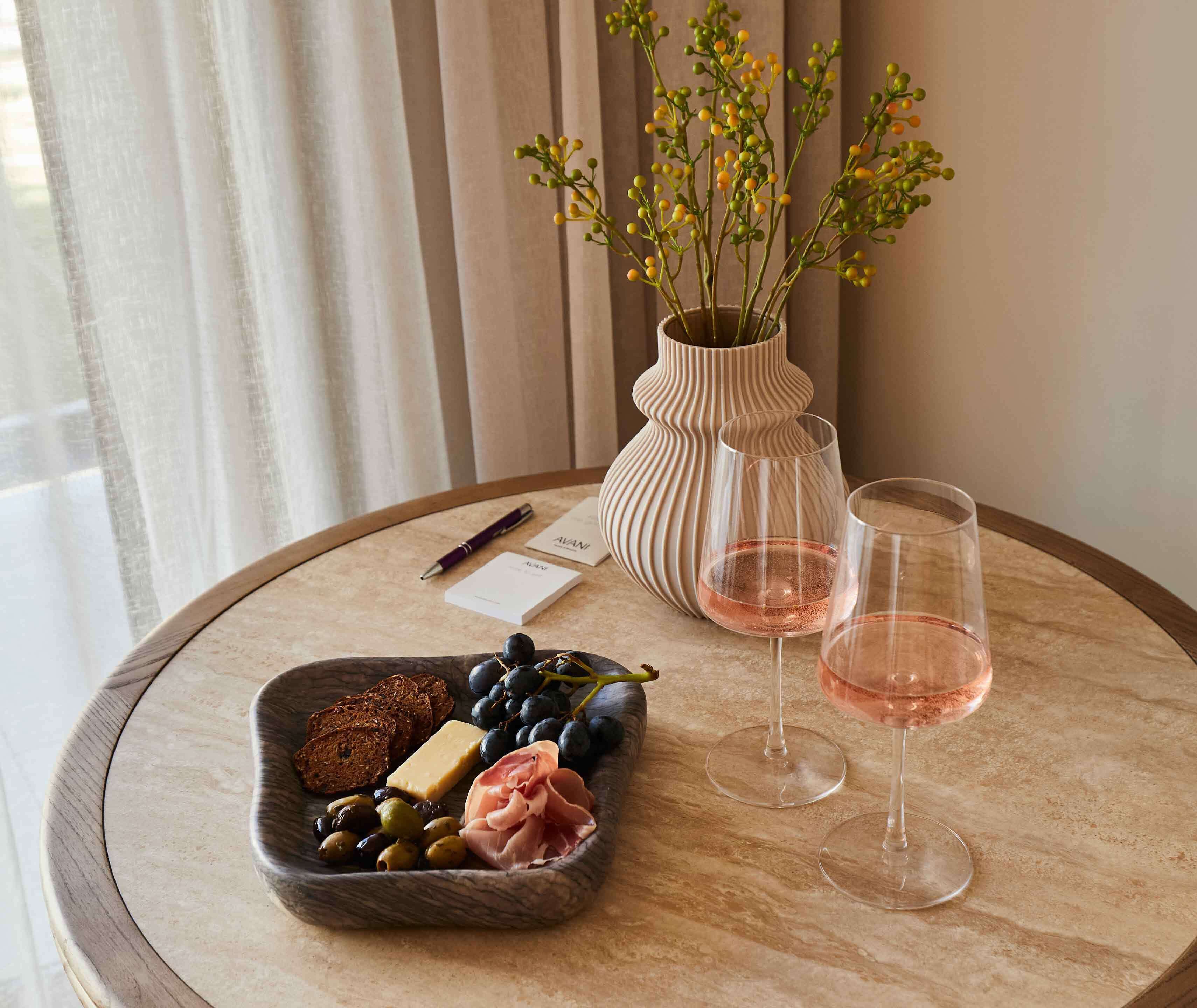 Une table avec une planche de fromages, des raisins, des olives et du jambon cru, deux verres de vin rosé, un vase de fleurs, un stylo et des cartes de visite.