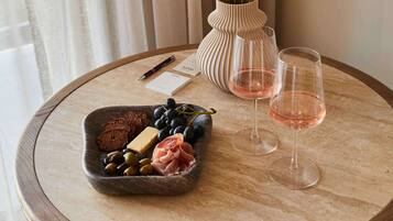 A table with a cheese board, grapes, olives, and prosciutto, two glasses of rosé wine, a vase with flowers, a pen, and business cards.